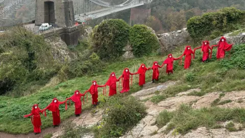 Red Rebel Brigade A line of women dressed from head to toe in red stand with the Clifton Suspension Bridge in the background. The group, known as the Red Rebel Brigade, appeared in various Bristol locations to highlight the start of COP 29 in Baku