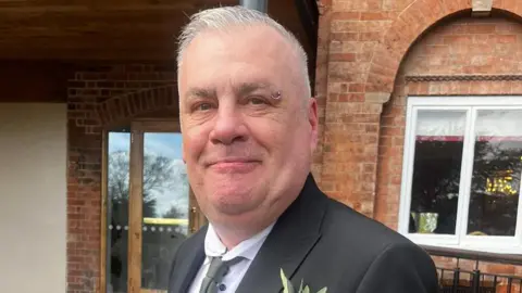 Sean is short cropped grey hair and a ring piercing on the end of the right eyebrow. He is wearing a black blazer over a white shirt and grey tie. There is a flower in the right breast pocket. He is standing in front of a redbrick building and smiling into the camera.