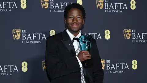 Getty Images David Jonsson stands in front of a hoarding with "EE Bafta Film Awards" printed on it. He's wearing a black suit with white shirt and bow tie.