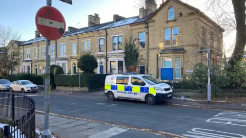 A police van parked at the end of a street on a T-junction with a red and white 'no entry' sign in the foreground on the other side of the same road. 