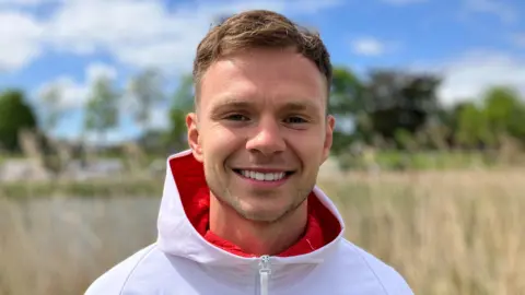 Lewis Adams/BBC Lewis Richardson has short blonde hair and is smiling in front of reeds by a pond. He is wearing a white hooded jacket.
