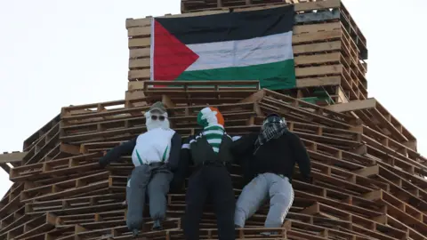 PA Media Three stuffed models are pinned to a bonfire, wearing tracksuits and various headwear - including a peaked caps and a green, white and orange balaclava. 