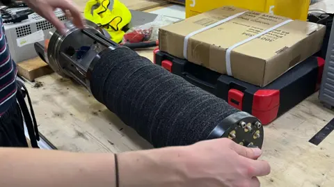 National Oceanography Centre A black hydrophone is laying horizontally on a worktop. It is shaped like a cylinder and a pair of hands are holding it at either end.