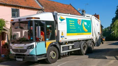 A waste collecting van with council staff sitting inside it wearing high-vis orange clothing. The van is white with blue and yellow council branding on it. There are cottages behind it. It is a sunny day with a blue sky above.