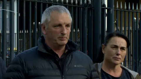 Michael McConville is standing looking slightly off camera with his lips pursed. He is wearing a black coat and black shirt. To his left a woman stands looking on. Behind them are black railings.