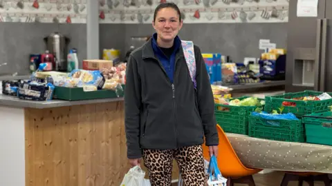 Leanne Hackett with dark hair tied back, wearing a black and blue fleece and leopard print leggings, holds two shopping bags as she stands in a kitchen full of boxes of food.