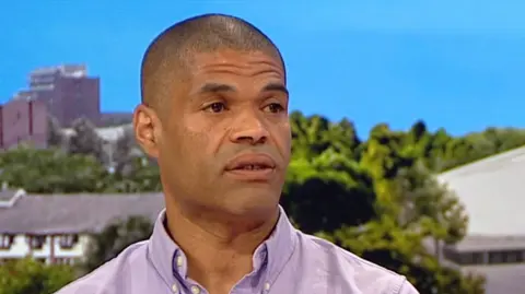 Man in a purple shirt sitting in a TV studio looking off camera. He is bald. 