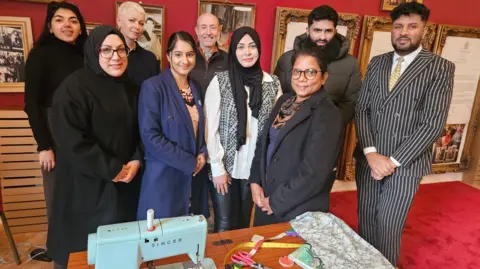 Aisha Iqbal/BBC A group of people of mixed ages and backgrounds standing behind a desk with a sewing machine and various tailoring and textile equipment and materials on it. They are in an ornately decorated room with framed vintage pictures on the deep red painted walls.