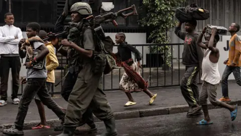 Getty Images A member of the M23 armed group walk alongside residents through a street of the Keshero neighbourhood in Goma, on January 27, 2025. 