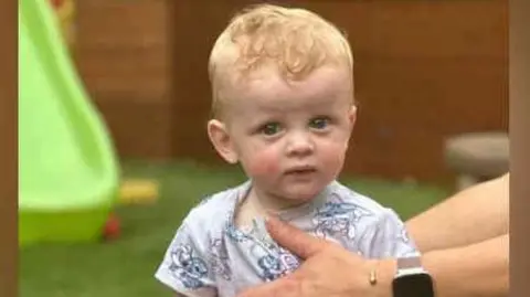 Oakley Barnett sitting on top of a large toy car in a garden. He is wearing a grey T-shirt and has blond hair.