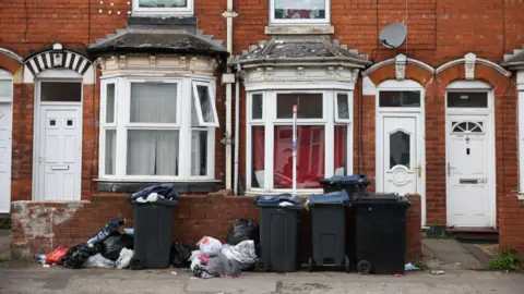 EPA/Shutterstock Several black bins, some with lids not fully down, with piles of rubbish near to them by a small wall. The wall is in front of terraced houses.
