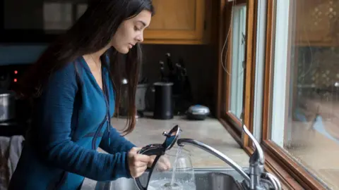 Getty Images Una mujer joven con cabello largo y oscuro llena una tetera transparente con agua en el fregadero de la cocina.