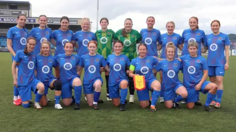 A team of female footballers pose together in two rows on a green pitch wearing a blue kit. The front row is on one knee, the back row is standing, and there ate two goalkeepers in the centre of the back row wearing a green kit.