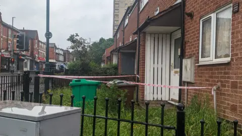 The front part of the house is with a red and white tape around it, the enlarged grass, and a green and brown eye