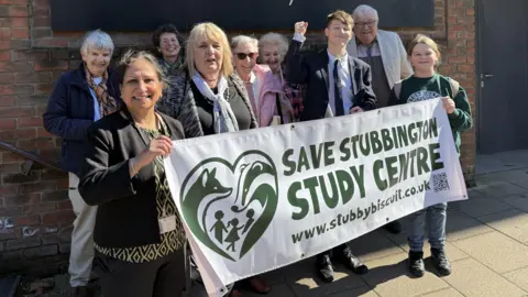 A group of people cheering and smiling holding a white banner with green writing on that says save stubbington study centre.