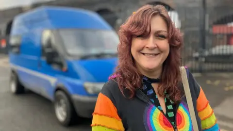 A woman with reddish-brown medium-length hair is smiling broadly in front of a large bright blue van in an industrial area of Leeds. She wears a colourful rainbow-striped cardigan and has a shoulder bag over one arm.