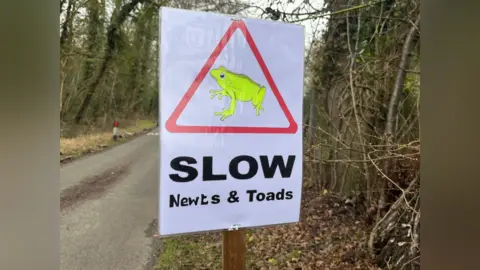 A white sign with a red triangle with a fluorescent yellow amphibian in the middle of it. The words 'Slow Newts & Toads' are written underneath.