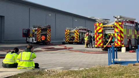 Three fire engines parked outside an industrial building, with two people sat in the foreground