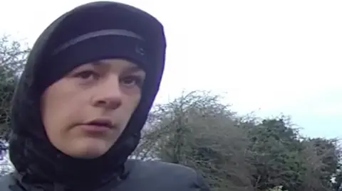 Body-worn camera footage of Kian Moulton released by West Midlands Police. The footage shows a young person wearing a black hoodie, hat and dark clothing outdoors. There are trees behind him in a park.