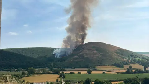 Tony Collier Photograph of a large grassy hill with the left side totally on fire. There is thick, black smoke pouring from the area. 