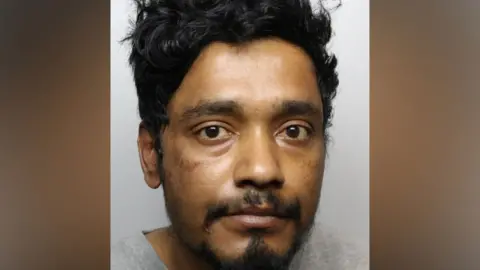 The image shows a close-up police mug shot of a man's head and upper shoulders against a plain, light grey background. He has medium-length, dark-coloured thick and slightly wavy hair.
