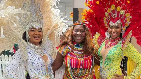 Three women including Bernadetta dressed in colourful Caribbean dance outfits. Bernadetta has her arms around both of them. The two women on either side both have sequin dresses and feathered headgear. The one on the left is in a white and blue outfit. The one on the right is in a yellow and emerald outfit, with a red headdress.
