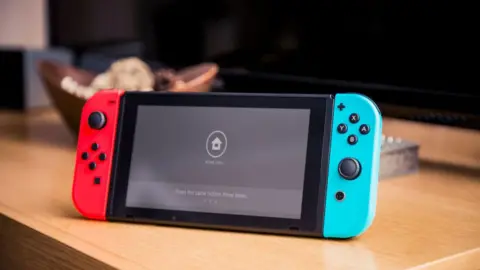 A Nintendo Switch console with red and blue joy con controllers attacked propper up on a desk. The screen is the home screen and says "press the same button three times".