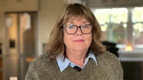 A bespectacled woman standing in front of a window. She is wearing a brown jumper and is looking at the camera.