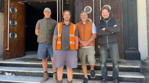 Simon Thake Four men in battered work gear stand proudly outside the steps to an entrance of a large building
