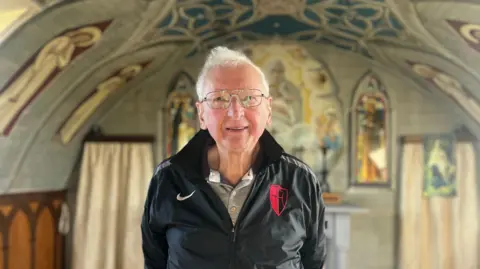 A man with white hair and glasses, standing inside the chapel. The alter can be seen in the background