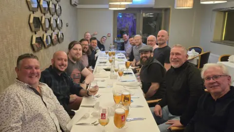 Man Up Durham A group of men are sat around a long table covered in a white table cloth smiling at the camera. A good number of them have pints in front of them.