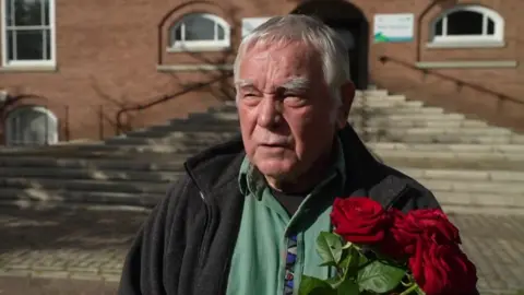 Mick Koch holding a bouquet of roses outside Devon County Council's main offices. He is wearing a grey fleece and a green shirt.