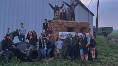Newborough Young Farmers A straw bale sculpture covered in young men and women. Some are standing on it, some are standing in front of it and others are sitting or standing to its side. Behind them is a grey barn and the back end of a trailer.