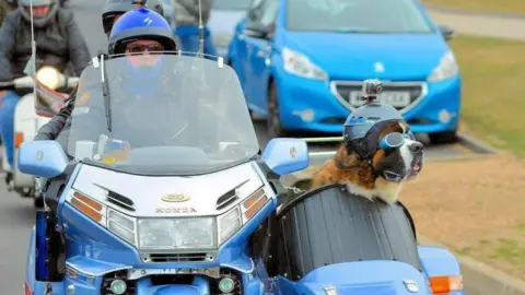 Alan Valkeith Alan Valkeith is riding a blue motorbike which has a sidecar with his St Bernard dog, Louis, sitting in it. Valkeith is wearing a black coat, black framed sunglasses and a blue helmet. Louis is also wearing a blue helmet and blue goggles.