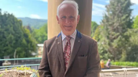 Man with short grey hair, brown tweed jacket stands on balcony with hillside backdrop