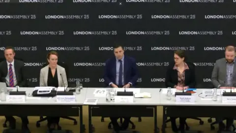 London Assembly Five panel members, including three men and two women, sit behind a row of desks at a meeting. Behind them is a backdrop that says London Assembly 25. 