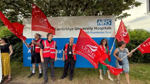 Steve Hubbard/BBC A group of people rallying for better pay in Cambridge. Most of them are wearing red high-visibility jackets bearing the logo of Unite the Union and several are waving large Unite the Union flags. They are pictured standing on a grass verge in front of a sign for Cambridge University Hospitals NHS Foundation Trust.