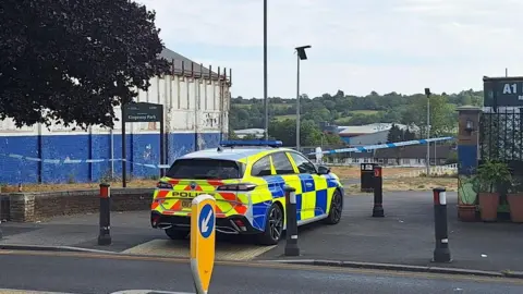 Bedfordshire Police A police car, in a road, between two buildings. There is police tape in front of the car, and bollards by a road. A tree is to the left and hills and trees are in the distance. Railings are to the right and a large path goes along a road.