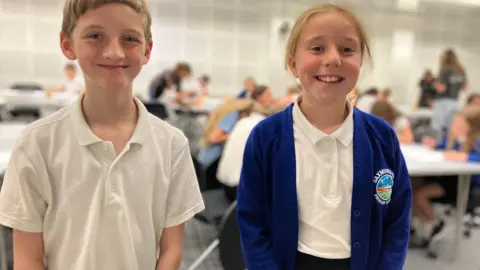 Jon Wright/BBC A 10-year-old boy and girl stand at the front of a classroom. they are in school uniform
