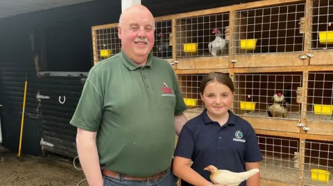 Alannah and her granda smiling at the camera as Alannah holds a baby rooster. Behind them are other roosters in seperate cages. Alannah's brown hair is tied up, she's wearing a navy polo shirt and her Granda is wearing a green polo shirt and has white hair with a moustache. 