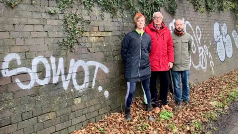 Three councillors stand in front of a wall with graffiti on it.