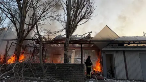 The State Emergency Service of Ukraine/X A firefighter stands next to a wall. Behind the wall is a destroyed building with a fire burning inside. 
