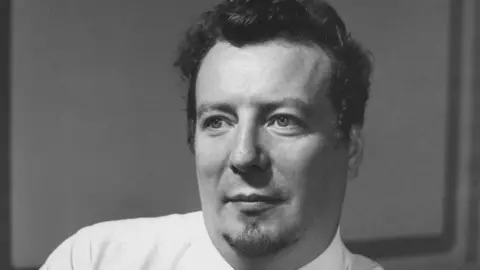 Stuart Burrows, shown in a black and white image, looks away from the camera wearing a shirt and tie. He has short dark hair and a goatee.