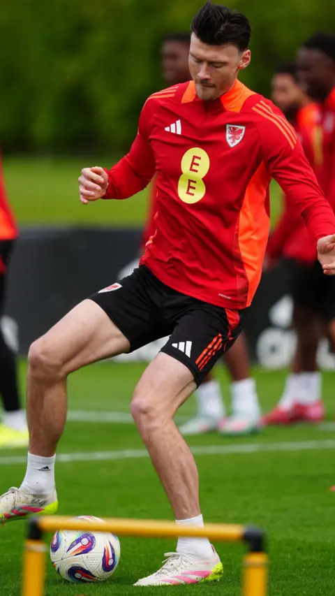 PA Kieffer Moore in training at Vale Resort, Hensol kicking a football wearing a Wales kit - with red shirt and black shorts. He is kicking a ball and there is a yellow small hurdle in front of him.