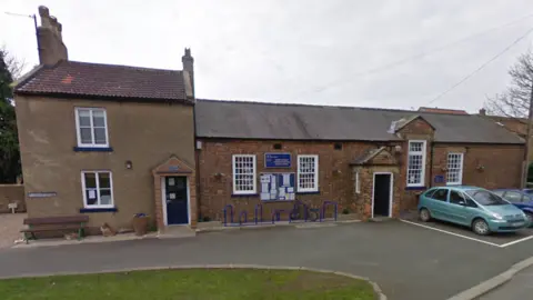 Google A small, one-storey red-brick school with a black door and white windows. There is a grey extension, which has two floors. Two cars are parked outside and there are blue bike racks below a notice board.