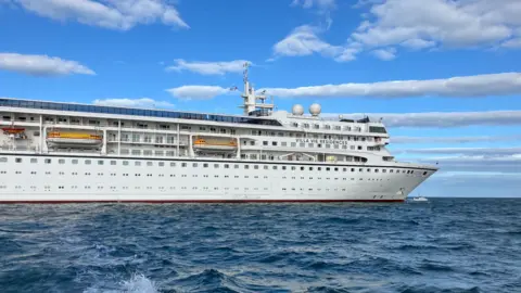 A wide shot of the Odyssey - a medium-sized white cruise ship sailing against a blue sky