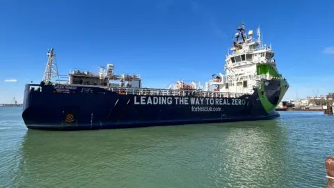 The Fortescue Green Pioneer vessel docked at the Port of Southampton. The words "Leading the way to real zero" are written on its side. It is a sunny day.