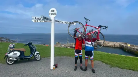 Supplied Eddie McGourley (l) and Stephen Foster celebrate as they arrive in John O'Groats. Eddie on  left wears a red cycling top and black leggings with red, white and blue detail. Stephen wears a red, white and blue cycling top and black cycling shorts with red, white and blue detail. They are holding aloft a red bike in front of a seaside harbour scene.