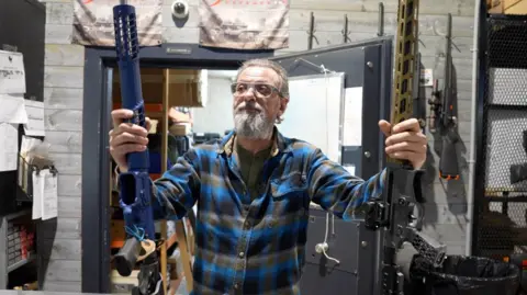 BBC Frank Nardi, bearded in a grey and blue plaid shirt and grey glasses, stands behind a counter propping up a long gun in each hand. 