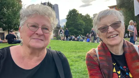 Mat Trewern/BBC Two women with grey hair sit on the grass. They both have glasses. The woman on the left is wearing black. The one on the right is wearing red orange and blue cardigan and a black T-shirt with a picture of sunflower and fish on it. 